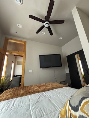 A bedroom with a bed, a ceiling fan, a television mounted on the wall, and a door leading to another room.