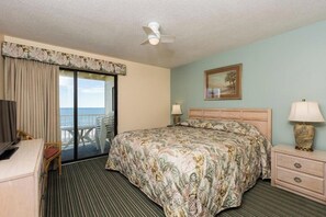 Habitación de hotel con cama, escritorio, silla, mesita de noche, lámpara, ventilador de techo, ventana con cortinas y un cuadro en la pared.