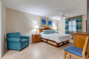 A bedroom with a bed, a chair, a nightstand, a desk, a ceiling fan, and two paintings on the wall. - Beachfront 2BR Retreat | Balcony + Full Kitchen (Cocoa Beach)