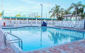 Una piscina con cerca blanca, sillas de descanso y palmeras al fondo.