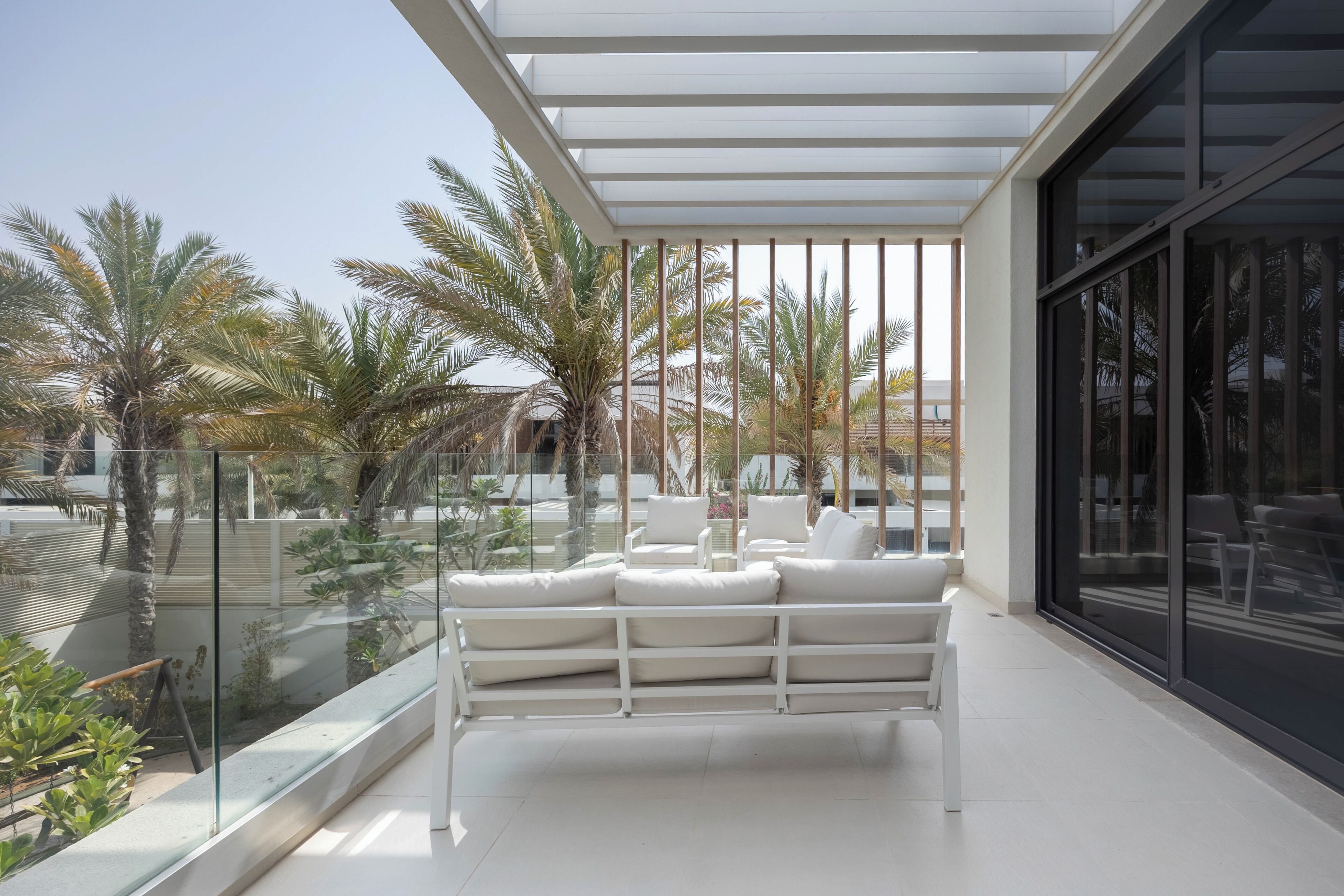 A modern outdoor seating area with white furniture, a glass balustrade, and a view of palm trees and buildings.