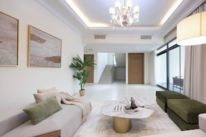 A spacious living room with a round coffee table, a sofa, and a green armchair. There is a large window on the right side, a staircase in the background, and a chandelier. - Bloomfields 5BR+ 2M Villa in West Yas (Abu Dhabi)
