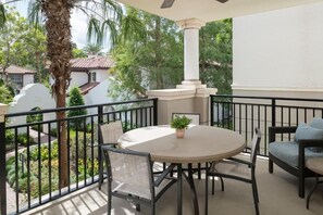 Een balkon met een ronde tafel en stoelen, een bloempot en uitzicht op een tuin en omliggende huizen.