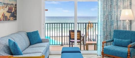 A room with a sofa, armchair, and a view of the beach through a sliding door.