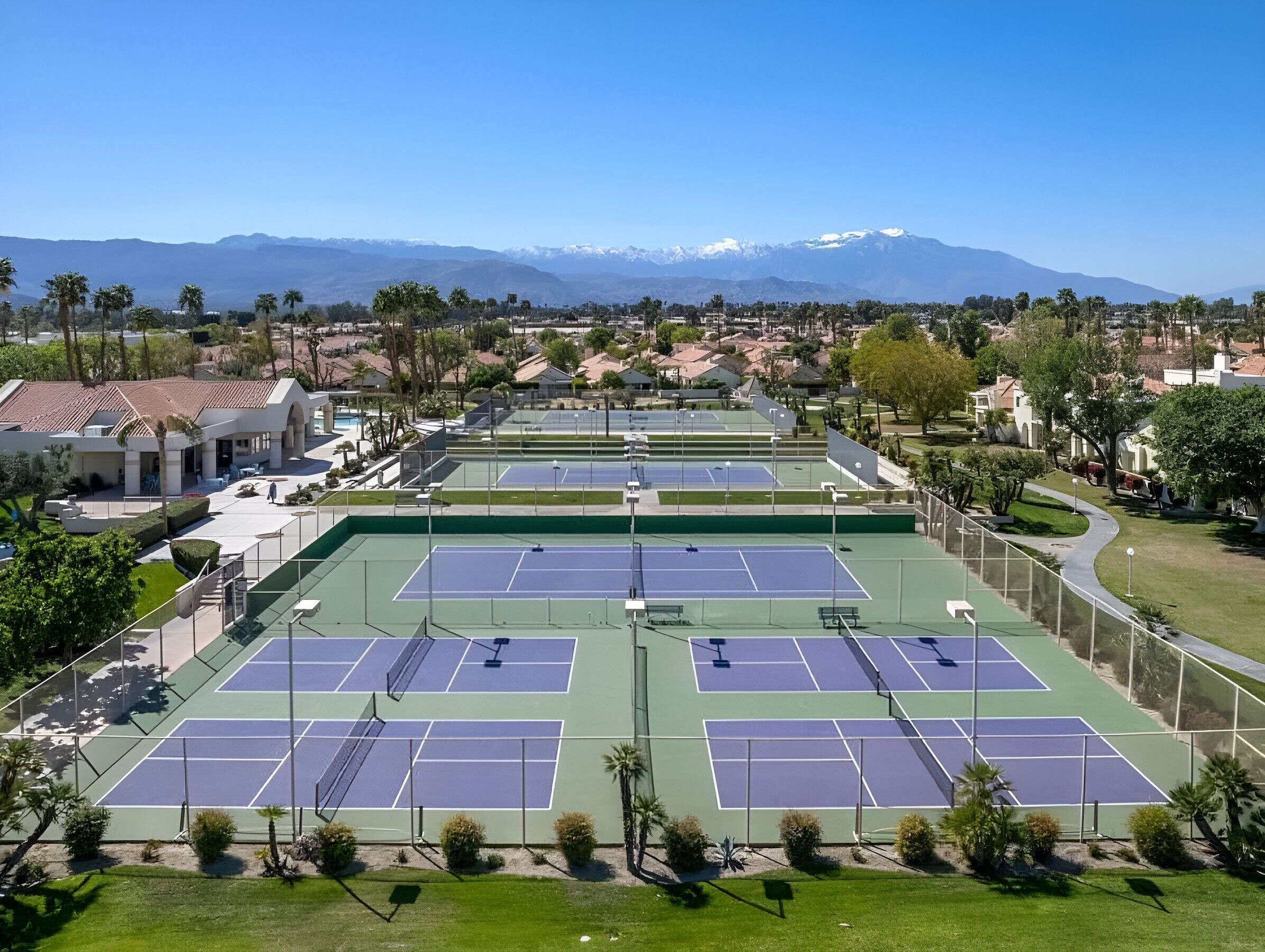 Un complexe de courts de tennis comprenant plusieurs terrains, entouré de bâtiments résidentiels et de palmiers, sous un ciel dégagé.