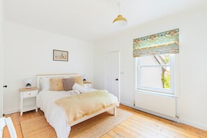 Ein Schlafzimmer mit Bett, Nachttischen, einem Fenster mit gemustertem Vorhang und einer Hängelampe. An der Wand hängt ein gerahmtes Bild.