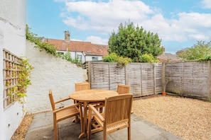 A patio with a wooden table and chairs, surrounded by a white wall and a wooden fence. - A comfortable two bedroom terrace house in NR2, sleeping four across two rooms, one with en suite shower-room. Free on street parking and log burner. (Norwich)