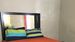 A single bed with a dark headboard and colorful pillows, a red mattress cover, and a plain wall with a subtle floral pattern.