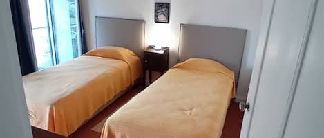 A room with two beds, a nightstand, a wall-mounted lamp, and a window with curtains.