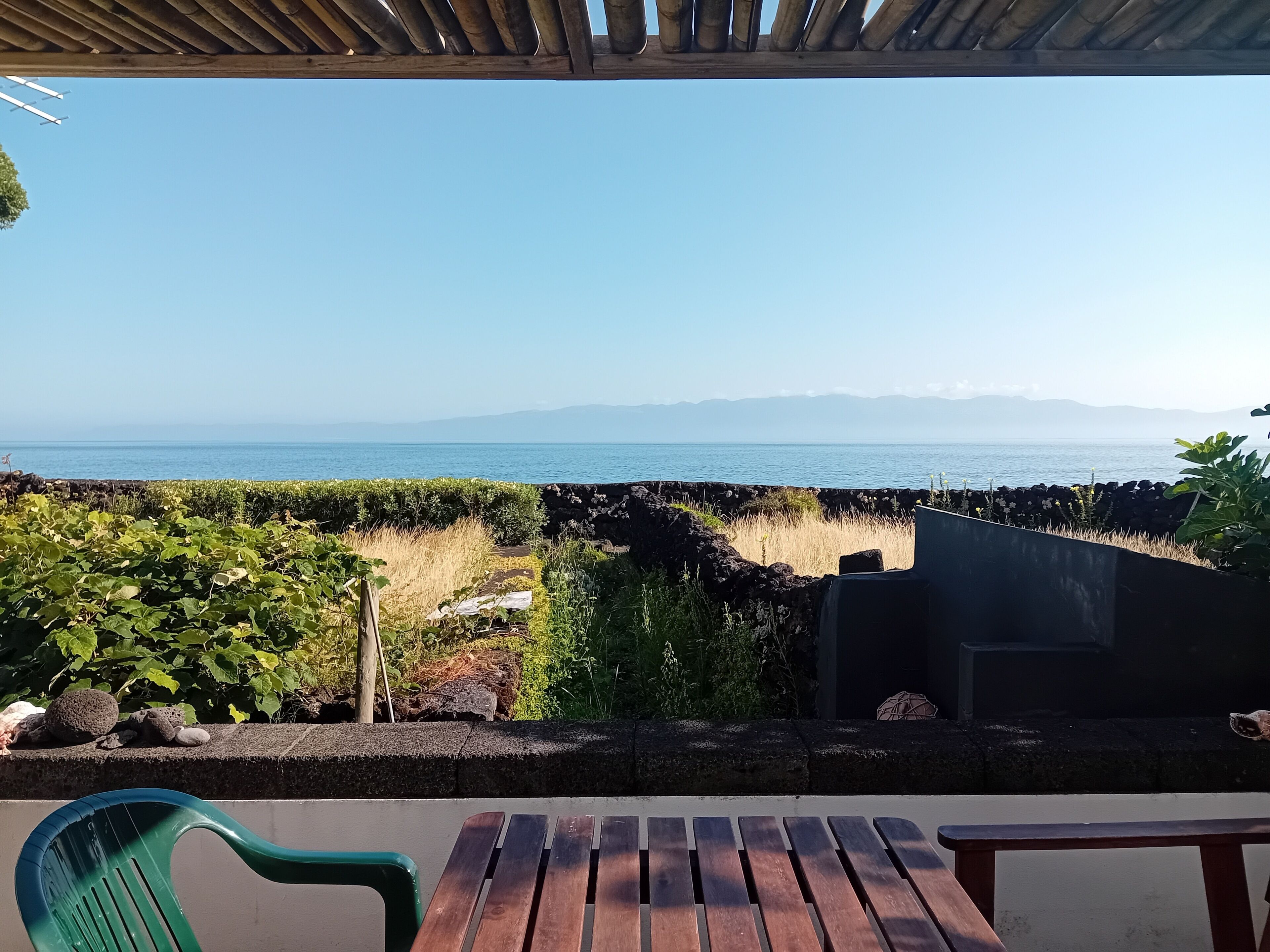 A wooden table and chairs on a balcony overlooking a coastal landscape with a clear view of the sea and distant mountains.