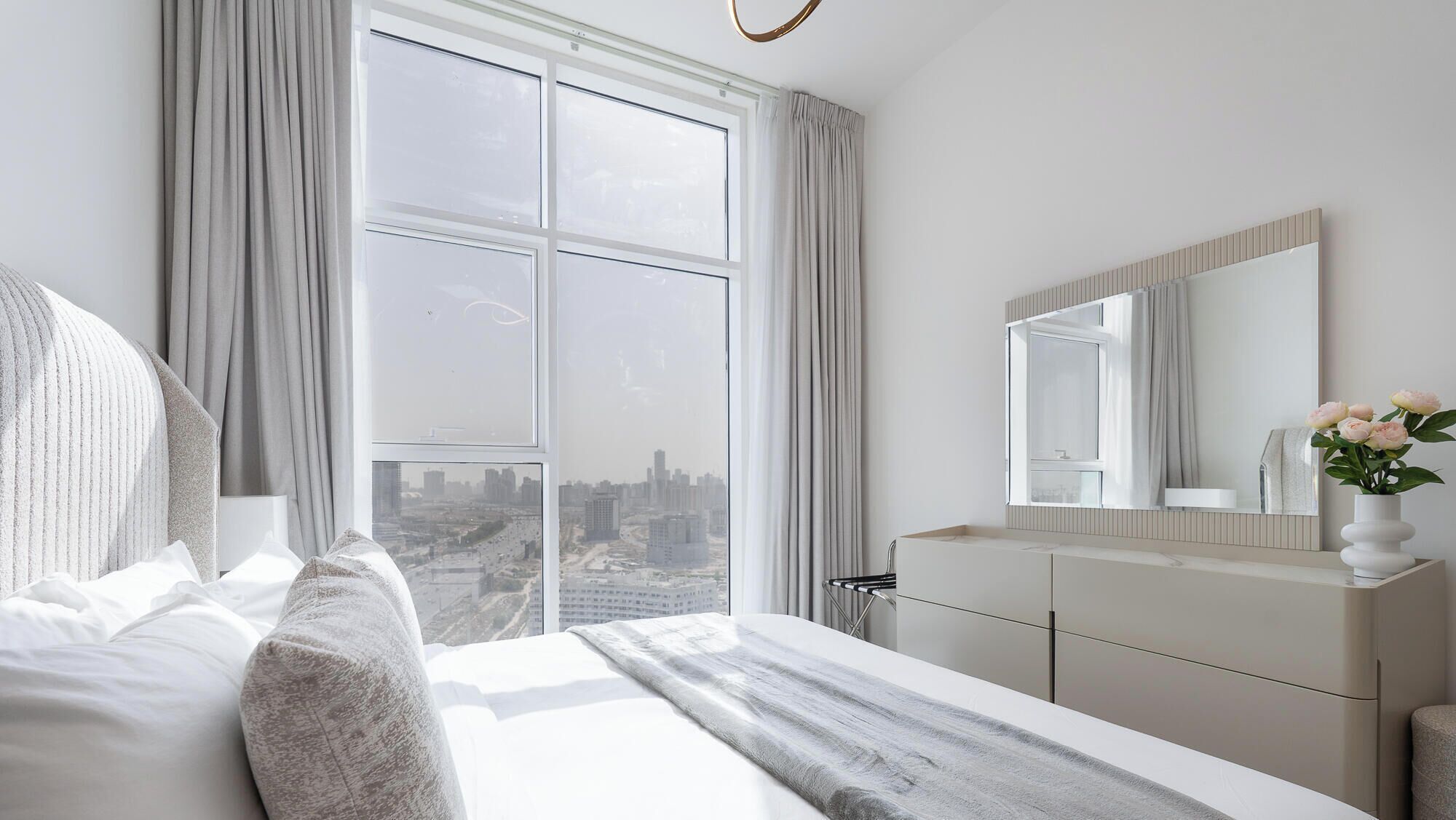 Une chambre moderne avec une grande fenêtre, un lit, une coiffeuse avec un miroir et un vase rempli de fleurs.