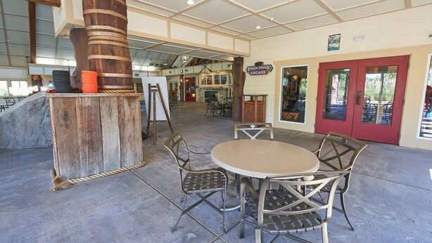 Ein Außenbereich mit einer Terrasse, einem runden Tisch und Stühlen, einer Holzsäule und einem Schild für eine Snackbar mit Spielautomaten.
