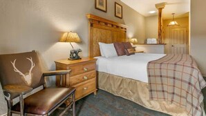 A hotel room with a bed, a wooden nightstand, a chair with a deer head, and a lamp.