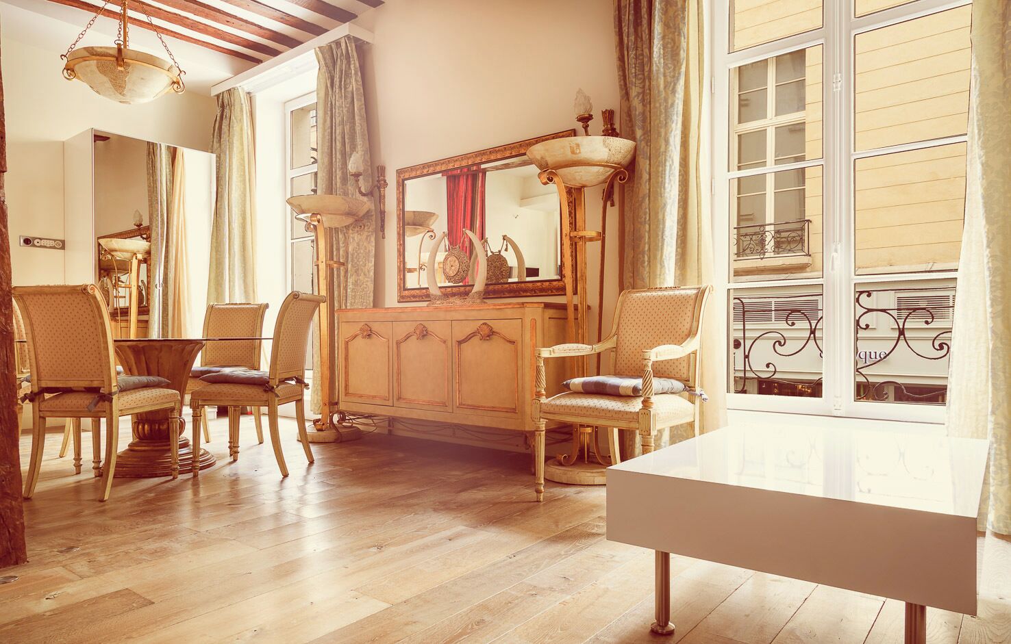 Een kamer met een houten vloer, een witte tafel en twee stoelen. Er is een spiegel met een decoratieve lijst en een lamp op een standaard.