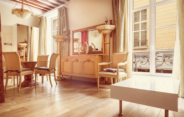 A room with a wooden floor, a white table, and two chairs. There is a mirror with a decorative frame and a lamp on a stand.