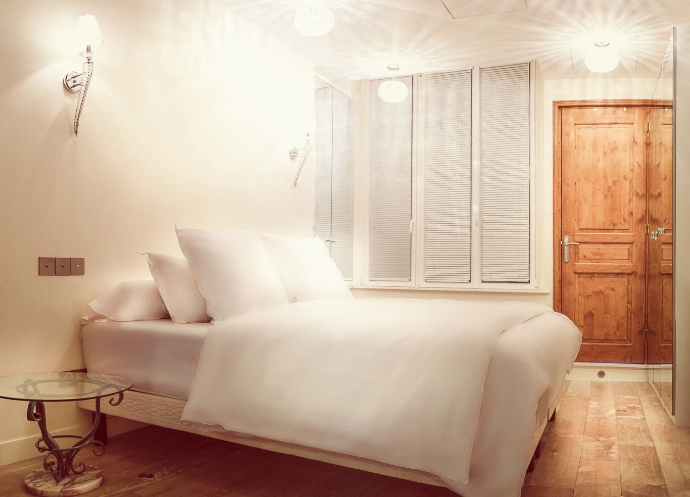 A bedroom with a bed, a glass table, a wooden door, and a window with blinds.