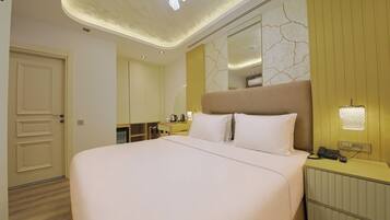 A hotel room with a large bed, a bedside table with a phone and charger, a mirror, and a small kitchenette area.