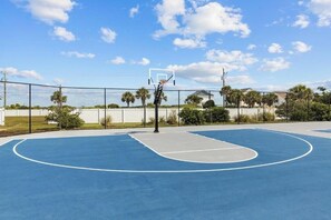 Una cancha de baloncesto al aire libre con aro, rodeada de vallas, palmeras y un edificio al fondo.