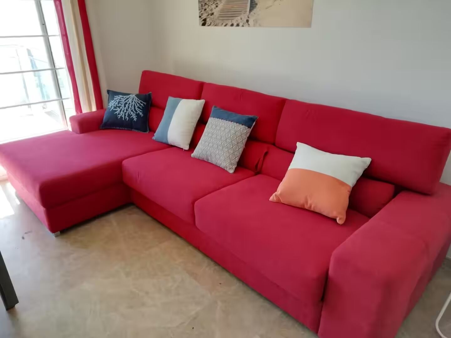A red sectional sofa with four pillows in a room with a tiled floor and a window with red curtains.