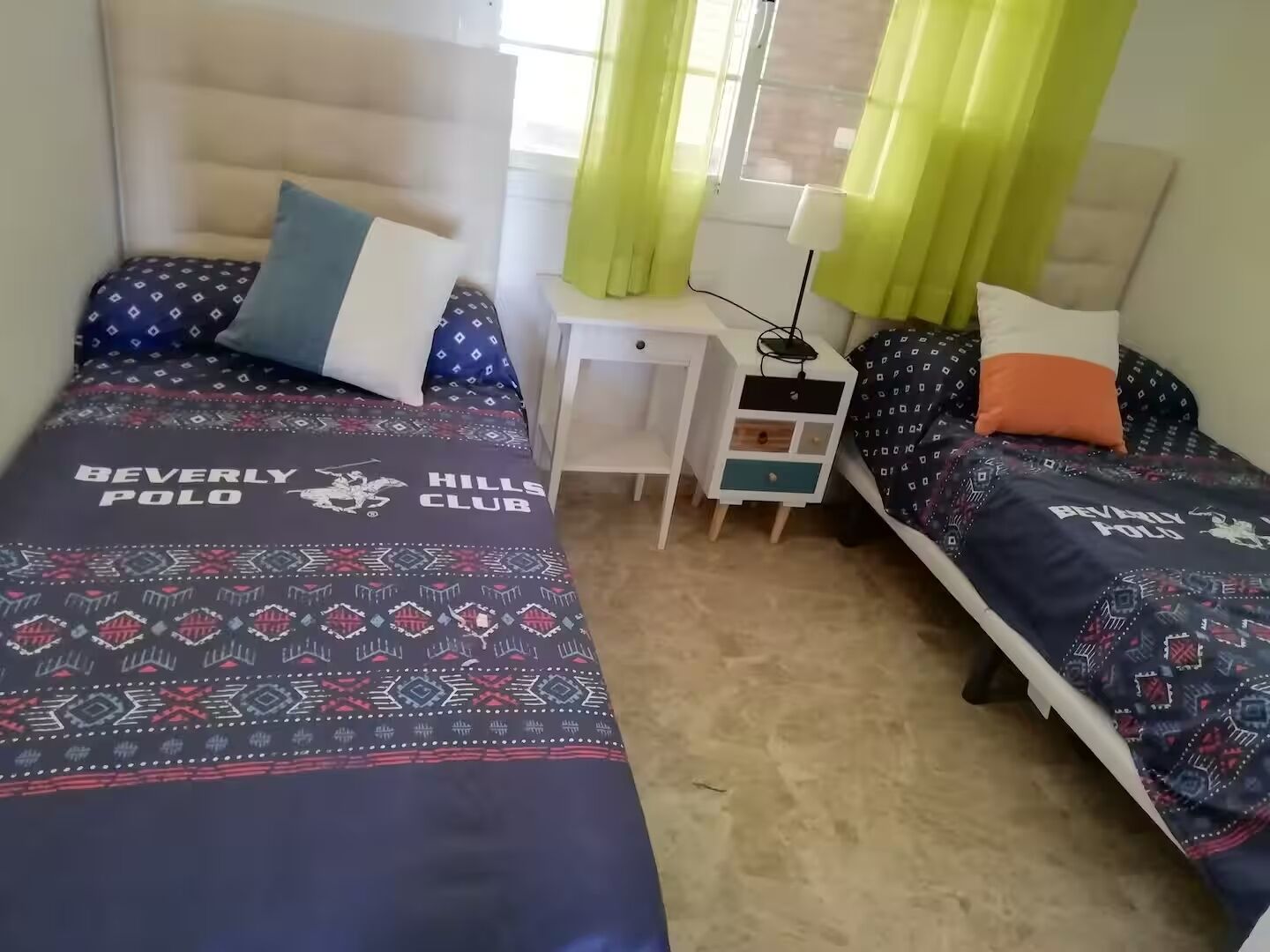 A bedroom with two beds, each with a patterned bedspread and pillows, a bedside table with a lamp, and a window with curtains.