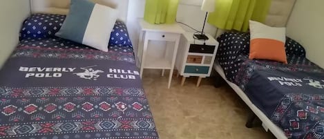 A bedroom with two beds, each with a patterned bedspread and pillows, a bedside table with a lamp, and a window with curtains.