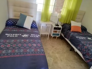 Ein Schlafzimmer mit zwei Betten, jeweils mit gemustertem Überwurf und Kissen, einem Nachttisch mit Lampe und einem Fenster mit Vorhängen.