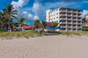 Un hotel sulla spiaggia con numerosi balconi, palme e una rete da pallavolo.
