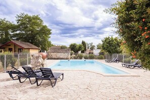 Una piscina con sillas de descanso, un patio de piedra y una zona vallada con árboles y un edificio al fondo.