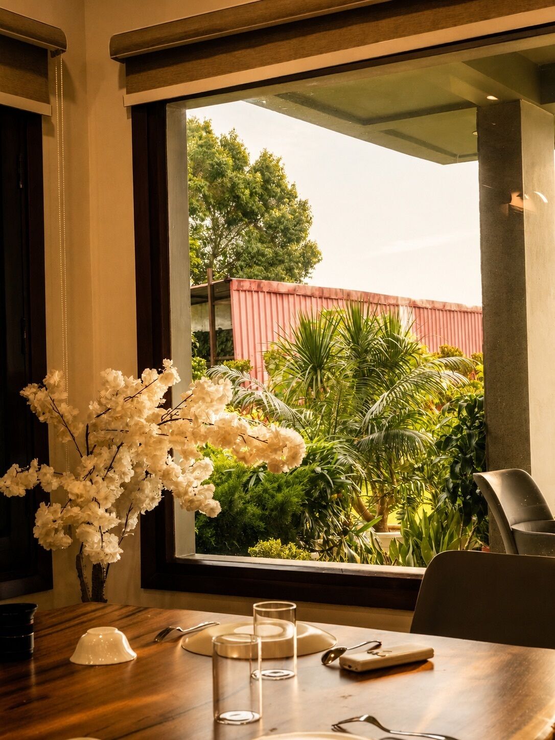 Un comedor con una mesa dispuesta para dos, un jarrón de flores blancas y vistas a follaje y un edificio rojo al exterior.