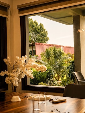 A dining area with a table set for two, a vase of white flowers, and a view of greenery and a red building outside.