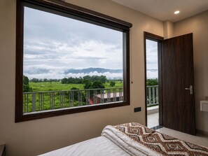 A bedroom with a large window offering a view of a green landscape and mountains.