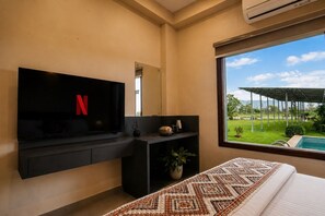 A hotel room with a large window, a flat-screen TV mounted on the wall, a bed with a patterned bedspread, and a small potted plant on the nightstand.