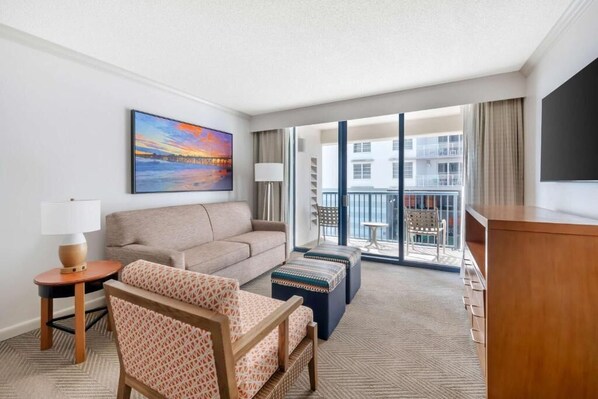 A hotel room with a sofa, armchair, ottoman, a painting, a TV, a lamp, and a balcony with a view. - 1 bed presidential at Daytona Regency (Daytona Beach)