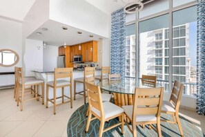 A modern kitchen with wooden cabinets and stainless steel appliances, a dining area with a glass table and wooden chairs, and large windows with a view of a building. - 1 bed presidential at Daytona Regency (Daytona Beach)