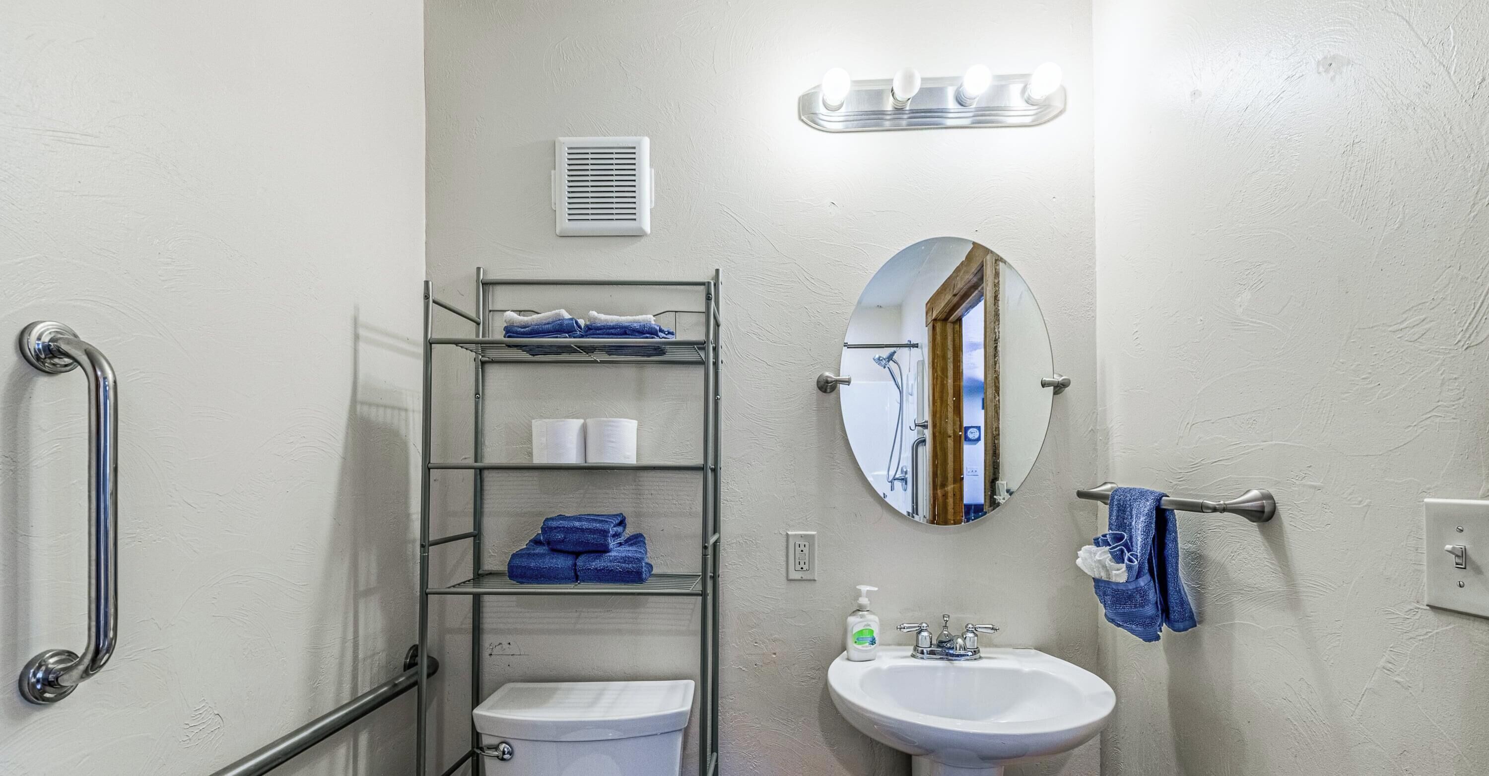A bathroom with a toilet, sink, mirror, towel rack, and a metal shelf unit with towels.