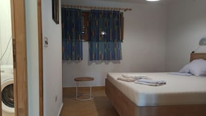 Una camera da letto con un letto, una finestra con tende, un comodino e una lavatrice nella stanza adiacente.