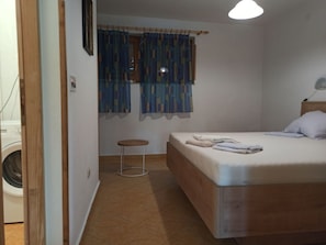 Ein Schlafzimmer mit Bett, einem Fenster mit Vorhängen, einem Nachttisch und einer Waschmaschine im Nebenraum.