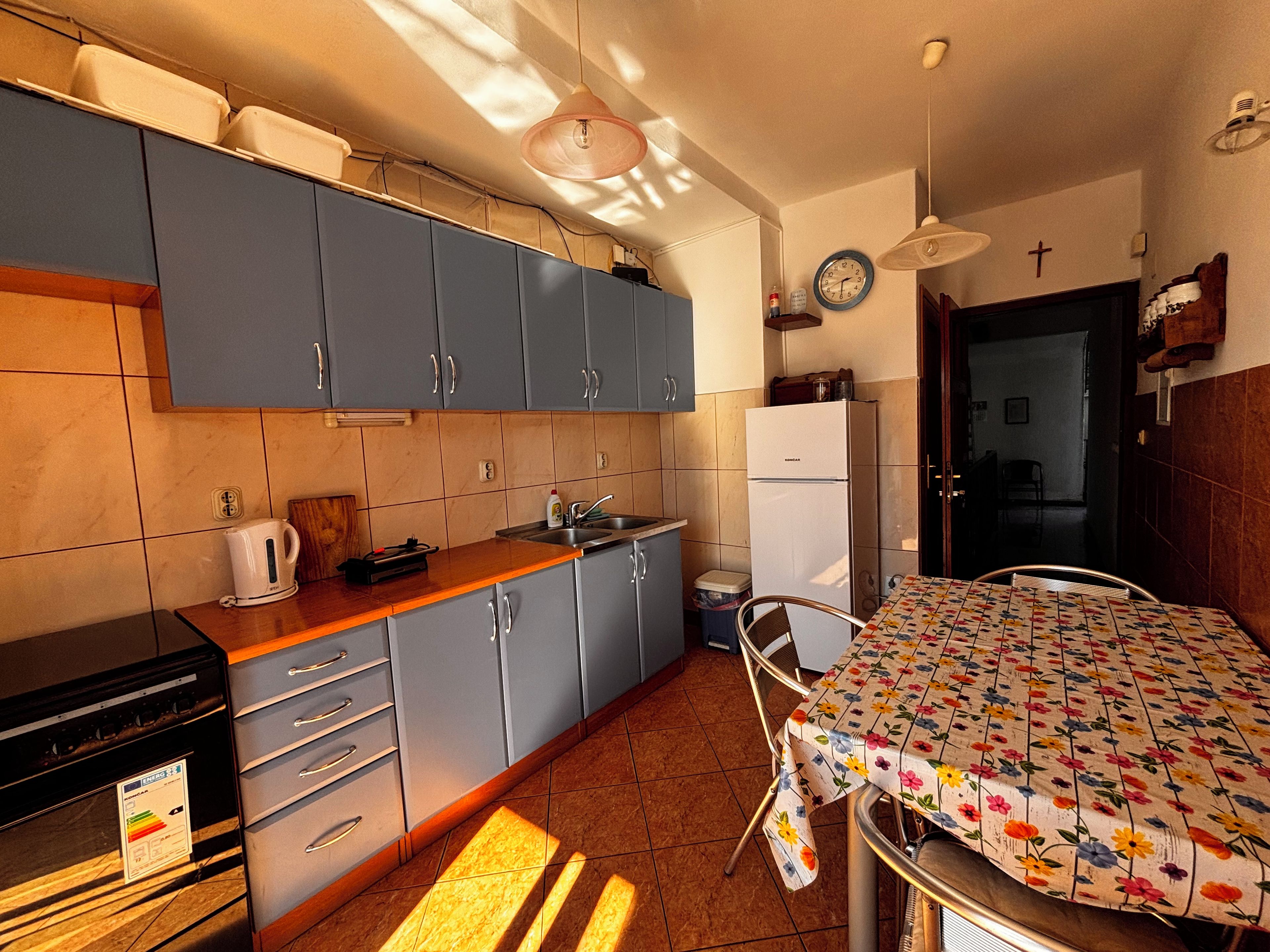 A compact kitchen with grey cabinets, a white refrigerator, a coffee maker, and a dining table with a floral tablecloth.