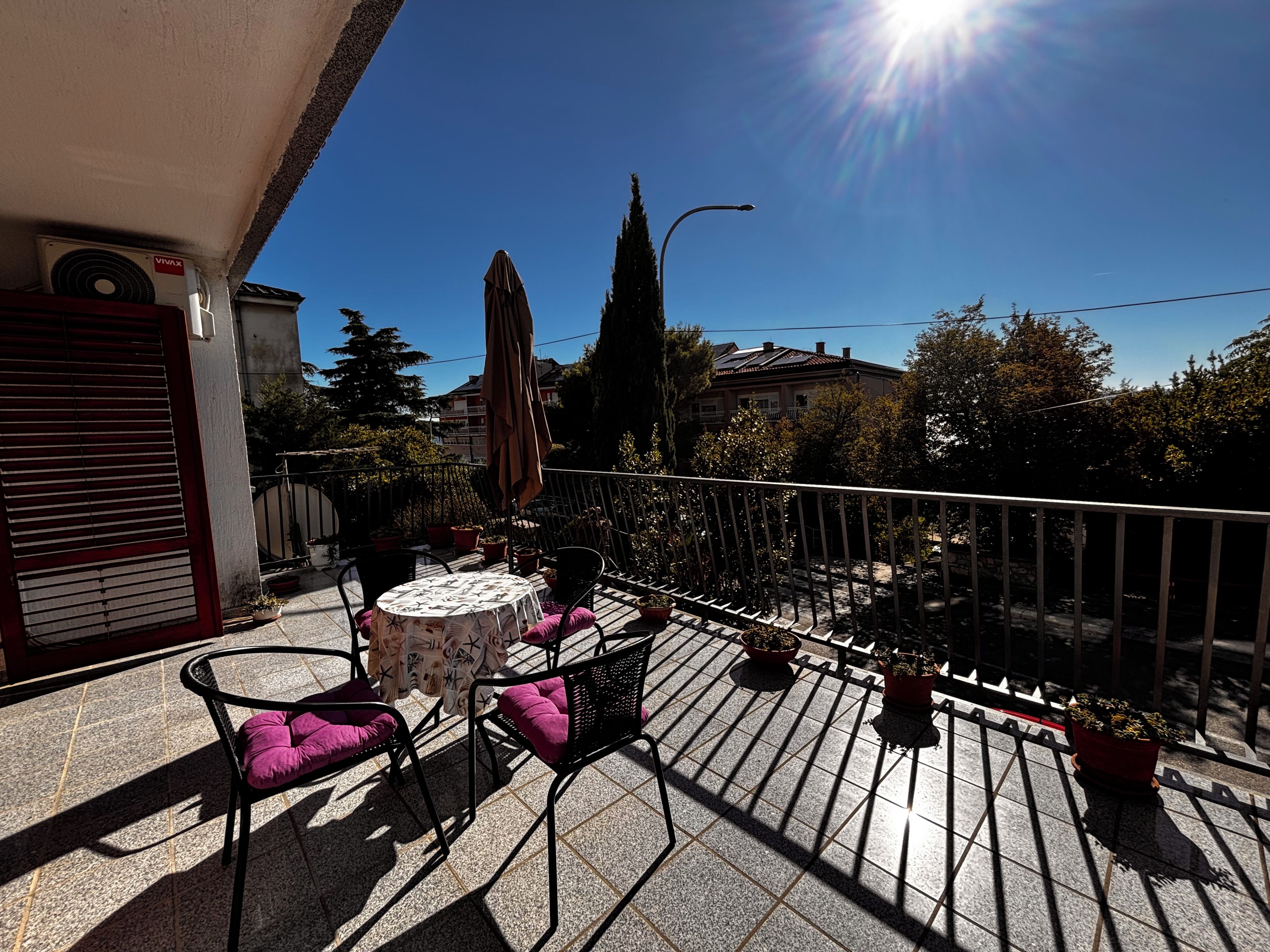 An outdoor balcony with a table and chairs, potted plants, and a view of the neighborhood.