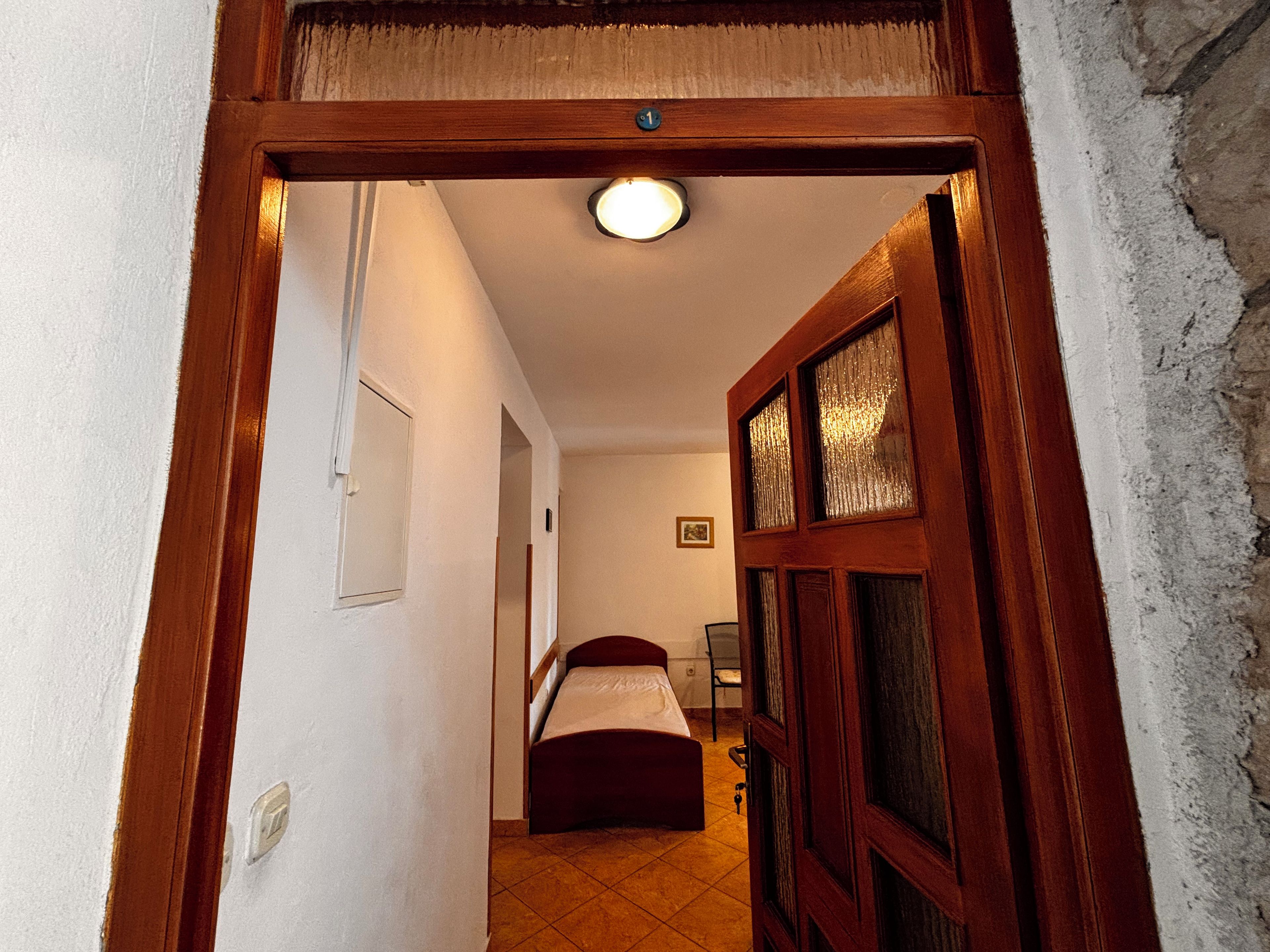 Corredor que leva a um quarto com uma cama, porta com placa de identificação e luminária no teto.