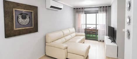 Una sala de estar moderna con un sofá beige, un aire acondicionado instalado, una obra de arte enmarcada, un televisor y un balcón con vistas.