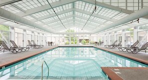 Ein großes Hallenbad mit klaren Glaswänden und -decke, umgeben von Liegestühlen und Tischen.