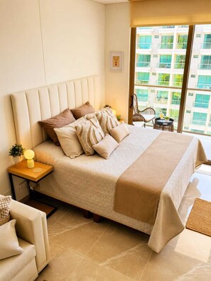 A hotel room with a large bed, a nightstand with a lamp, a chair, and a view of a building through the window. - Luxury Sky Retreat / Rooftop with Ocean View (Santa Marta City)
