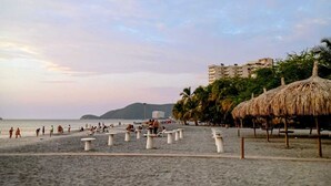 Ein Strand mit Strohdächern, ein Sandbereich mit Tischen und ein Gebäude im Hintergrund.