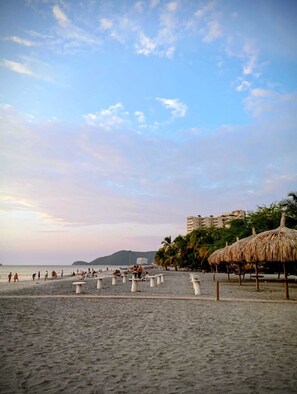 Ein Strand mit Strohdächern, ein Sandbereich mit Tischen und ein Gebäude im Hintergrund.