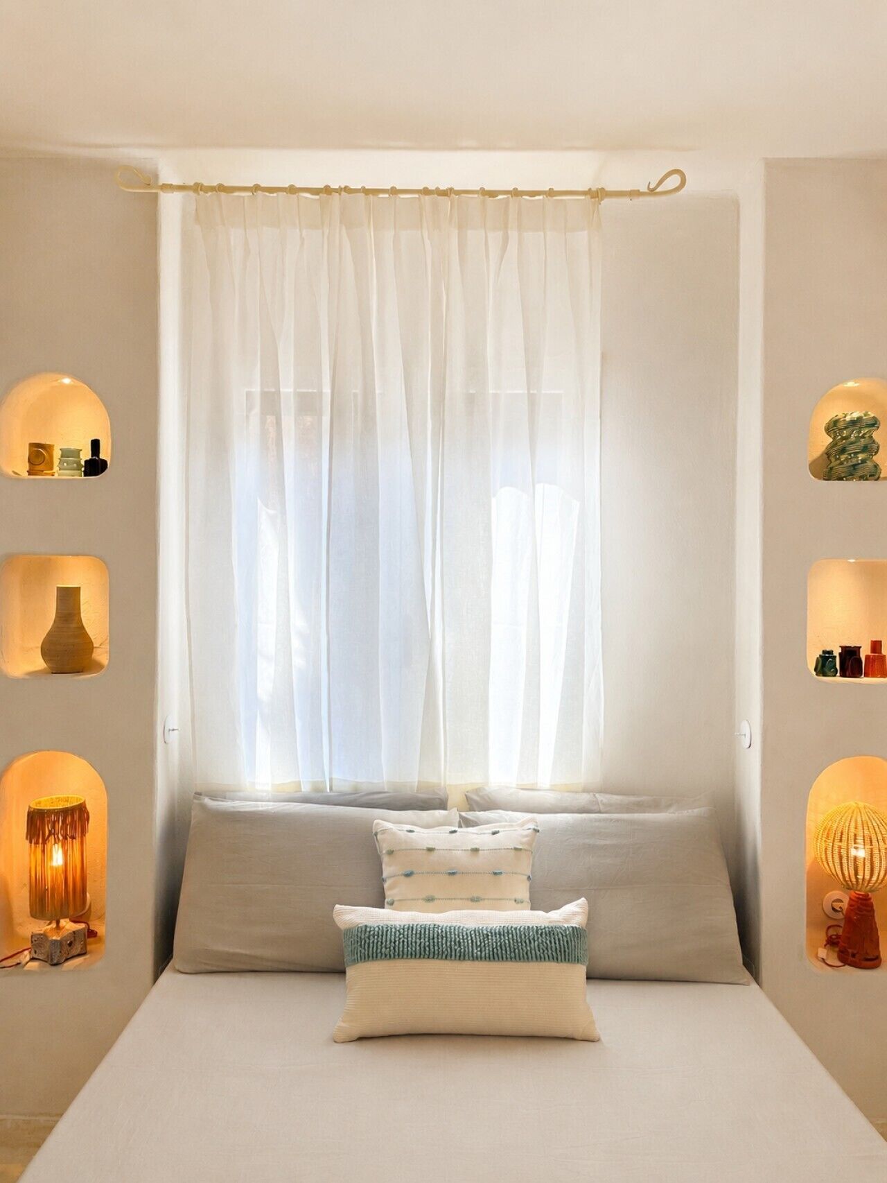 A bedroom with a bed, a window with sheer curtains, and wall-mounted shelves with various items.