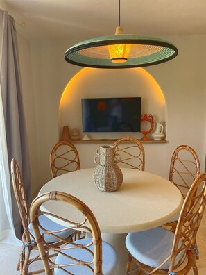 A round dining table with six wicker chairs, a woven vase, and a television mounted on a wall with an arched alcove. - Sunset Suite Sardinia 1.5 (Porto Rotondo)