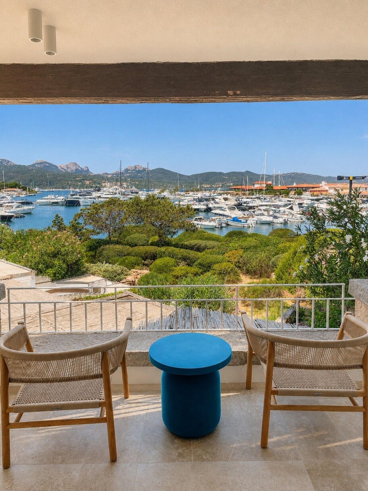 Un balcon agrémenté de chaises en osier et d’une table bleue, donnant sur une marina où l’on peut admirer des bateaux et une végétation luxuriante.