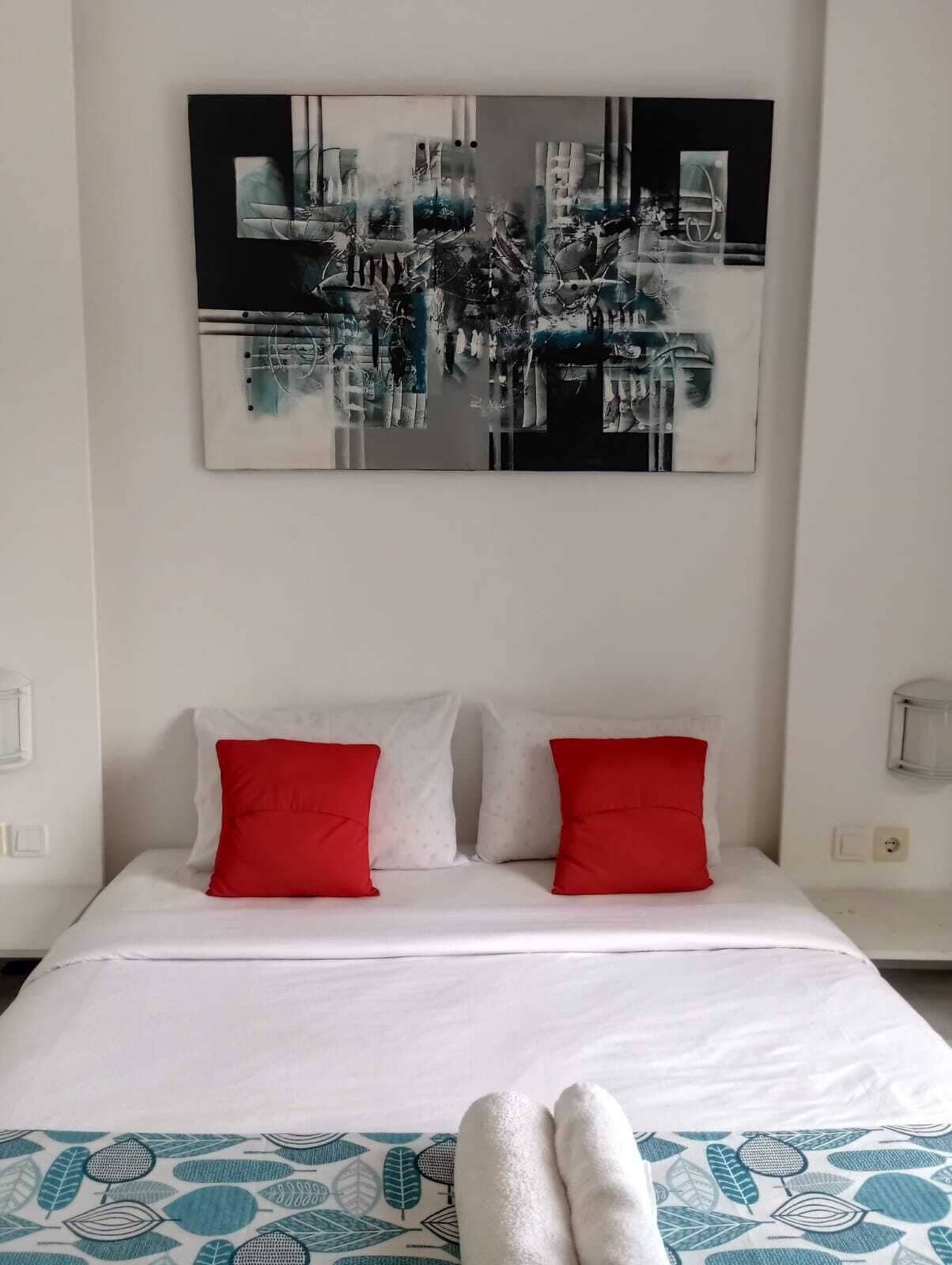 A bed with white bedding and two red pillows, a large abstract painting above it, and a patterned throw at the foot of the bed.