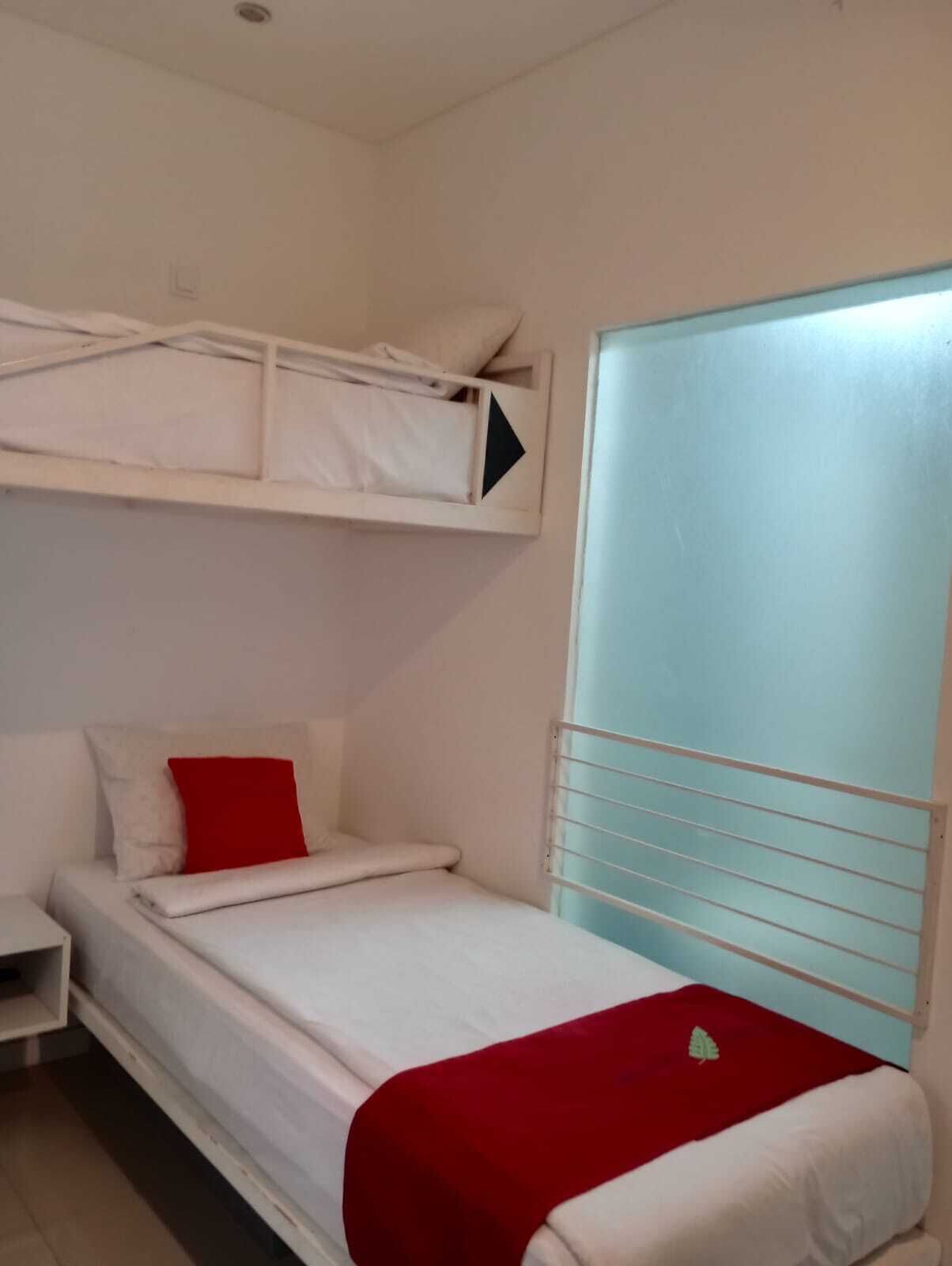 A single bed with a red pillow and a white blanket, a white nightstand, a white wardrobe with a frosted glass door, and a white ceiling with a light fixture.